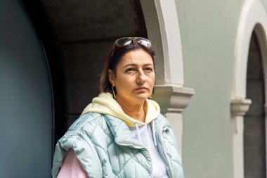 Kış kıyafetleri içinde düşünceli bir kadın kemerli bir duvarın yanında duruyor.