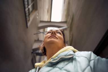 Şehir perspektifi: Kabarık ceketli kadın dar sokakta yukarı bakıyor.