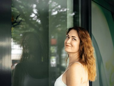 Dalgalı saçlı, gülümseyen kadın modern cam pencereye yansıyor.