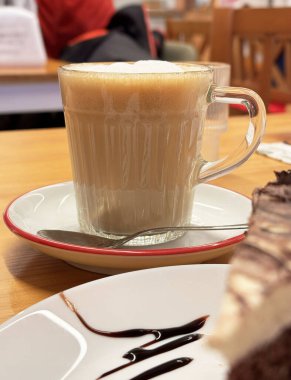 Kafe masasında cam bardakta kremalı kapuçino ve çikolatalı pasta dilimi..