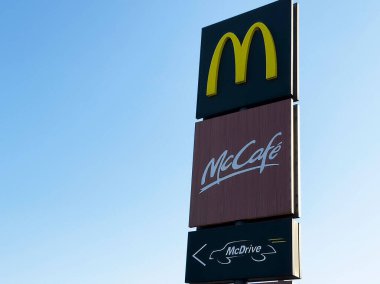 McDonald 's' ın simgesel tabelası McCafe 'yi ve açık mavi gökyüzüne karşı arabalı servis seçeneklerini gösteriyor, fast food kültürünü ve konseptini temsil ediyor.