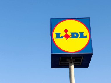 Açık mavi gökyüzüne karşı Lidl süpermarket işareti canlı renkler ve markalar sergiliyor, indirimli perakende alışveriş kavramını temsil ediyor