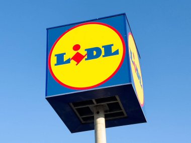 Lidl süpermarket tabelası açık mavi gökyüzü karşısında canlı renkler ve markalar sergiliyor, perakende alışveriş deneyimi ve kolaylık kavramını temsil ediyor