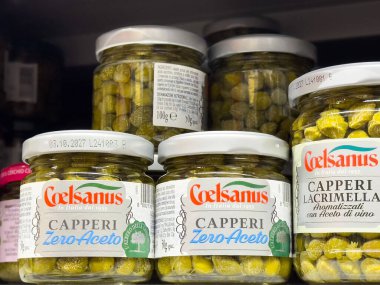 Bir süpermarket rafında sergilenen kapariler çeşitli marka ve türleri gösteriyor, canlı yeşil rengi vurguluyor ve eşsiz ambalajlar, sağlıklı beslenme konsepti