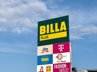 BILLA PLUS süpermarket işareti açık mavi gökyüzüne karşı çeşitli marka ve logoları sergiliyor, alışveriş kolaylığı ve perakende çeşitliliği kavramını temsil ediyor