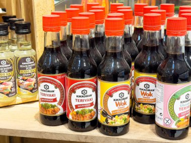Bir marketin ahşap rafında çeşitli aromalar ve sağlıklı pişirme seçenekleri sunan çeşitli teriyaki sosu ve tava sosu şişeleri sergileniyor.