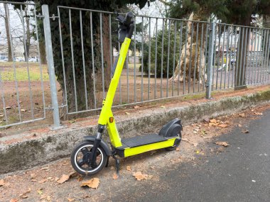 Parlak sarı elektrikli scooter şehir kaldırımına park edilmiş, yaprakları dökülmüş..
