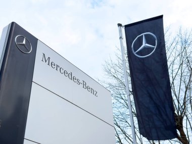 Bulutlu bir günde yapraksız ağaçların yanında işaret ve bayrak üzerine Mercedes Benz logosu.