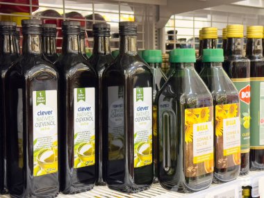 Yeşil ve siyah ambalajlı çeşitli zeytinyağı şişeleriyle market rafı..