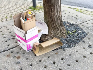 Kaldırımın köşesinde çakıl taşları ve beton deliklerle dolu karton kutular ve ağaç var..