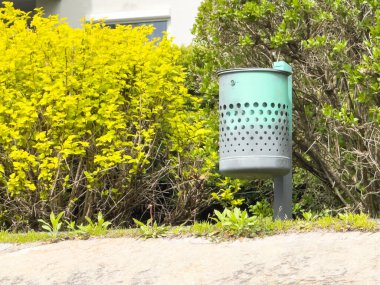 Yeşil parktaki çalıları ve sarı yaprakları olan açık hava metal çöp kutusu..