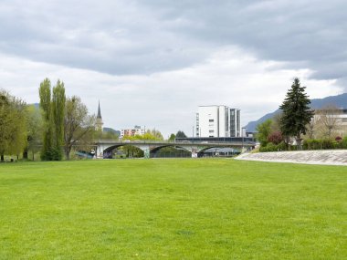 Villach, Avusturya - 17 Nisan 2025: Köprüsü olan yeşil park ve bulutlu bir gökyüzünün altındaki binalar kentsel doğa manzarasını gözler önüne seriyor