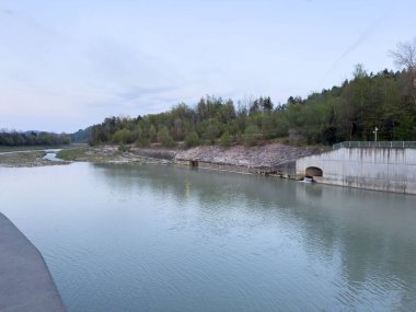 Orman arka planında huzurlu nehir kenarı ve alacakaranlıkta beton yapı.