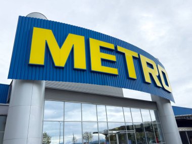 Bulutlu gökyüzüne karşı mavi ve sarı işaretli Metro süpermarketi girişi.
