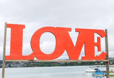 Kırmızı ahşap aşk işareti bulutlu gökyüzü ve teknelerle sakin göl kenarlarında.