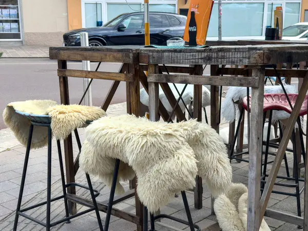 Ahşap masalar ve kaldırımda koyun postu kaplı bar tabureleri olan rahat bir açık hava koltuğu.