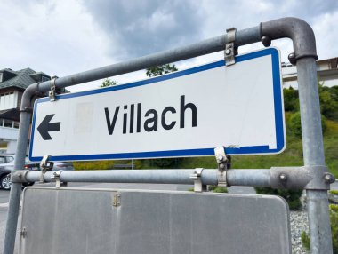 Bulutlu gökyüzü ve şehir arkaplanı olan Villach yönlü sokak tabelası.