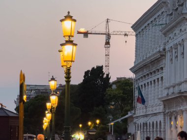 Venedik, İtalya - 23 Haziran 2025: Akşam sokak sahnesinde parlayan lambalar, tarihi binalar ve canlı bir kentsel atmosfer yaratan bir inşaat vinci yer alıyor.