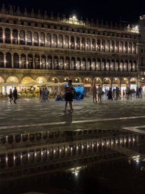 Venedik, İtalya - 23 Haziran 2025: Geceleri Venedik 'teki St. Mark Meydanı' nda aydınlanan binaların güzel yansımaları ve atmosferin keyfini çıkaran insanlar