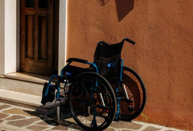 Burano, İtalya - 25 Haziran 2025: Tekerlekli sandalye turuncu duvarlı ahşap kapının yanında duruyor, ulaşılabilirliği ve günlük faaliyetlerdeki hareketliliğin önemini vurguluyor