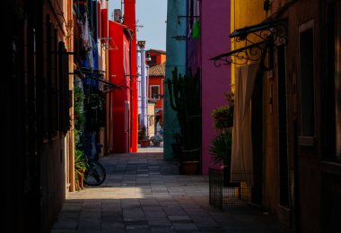 Burano, İtalya - 25 Haziran 2025: Burano, İtalya 'da dar bir sokak, renkli binalar ve yeşillikler içeriyor, büyüleyici bir şehir atmosferi yaratıyor.