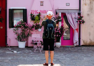 Burano, İtalya - 25 Haziran 2025: Sırt çantalı bir adam renkli çiçeklerle çevrili güzel pembe bir binanın görüntüsünü yakalıyor, canlı bir sahne yaratıyor.