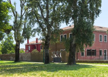 Burano, İtalya - 25 Haziran 2025: Güneşli bir yerleşim bölgesinde ağaçlarla çevrili geleneksel İtalyan binaları, pitoresk bir atmosfer yaratıyor.