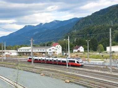 Villach, Avusturya - 10 Temmuz 2025: Dağlar ve yeşilliklerle çevrili modern tren raylarda ilerliyor, ulaşım ve doğanın uyumu vurgulanıyor