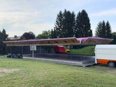 Villach, Avusturya - 20 Temmuz 2025: Renkli bir karnaval biniş yapısı, arka planda ağaçlarla birlikte çimenli bir alana park edilmiş büyük bir tepe örtüsüne sahip, neşeli bir ortam yaratıyor.