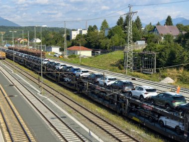 Arnoldstein, Avusturya - 20 Temmuz 2025: Yeni vagonlar taşıyan yük treni, yeşil tepeleri ve dağları olan düz raylı vagonlara istiflenmiştir.