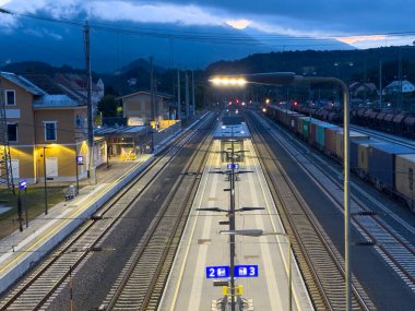 Arnoldstein, Avusturya - 24 Temmuz 2025: Aydınlatılmış platformlar ve kargo konteynerlerinin yer aldığı tren istasyonu sahnesi, demiryolu altyapısı ve ulaşım unsurlarının altı çizili