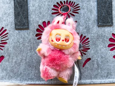 Villach, Avusturya - 28 Temmuz 2025: Kırmızı gözlü ve neşeli gülüşlü sevimli pembe pelüş oyuncak, çiçek desenli yumuşak gri arka planda asılı