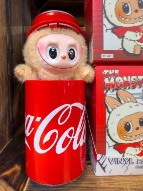 Villach, Avusturya - 30 Temmuz 2025: Pelüş oyuncak karakteri, arka planda koleksiyon kutuları ile eğlenceli bir ortam yaratarak, Coca-Cola kutusundan eğlenceli bir şekilde ortaya çıkıyor