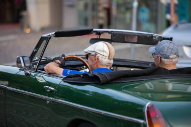 Velden, Avusturya - 18 Haziran 2025: Klasik yeşil üstü açık spor araba yaşlılar tarafından canlı renklerle dolu bir şehir sokağına sürülüyor