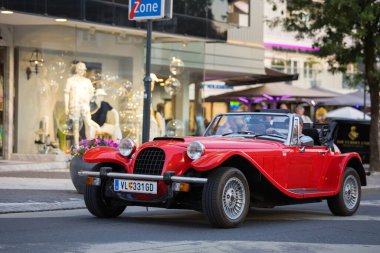 Velden, Avusturya - 18 Haziran 2025: Arka planda lüks sürüş ruhunu yakalayan modaya uygun dükkanlarla şehir caddesinde dolaşan klasik kırmızı spor araba