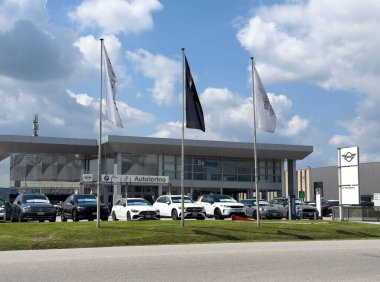 Udine, İtalya - 26 Ağustos 2025: Birden fazla araç, bayrak ve parlak mavi gökyüzü altında çağdaş bir binanın yer aldığı bir araba galerisinin dış görünümü