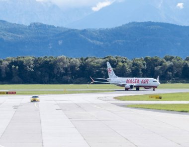Klagenfurt, Avusturya - 25 Eylül 2025: Malta Hava Yolları yeşil ağaçlar ve dağlarla çevrili havalimanında uçuyor, seyahat atmosferini yakalıyor