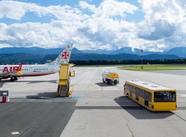 Klagenfurt, Avusturya - 25 Eylül 2025: Pistte uçak, sarı otobüs ve yakıt kamyonlu havaalanı manzarası, seyahat lojistiği ve manzaralı dağları vurguluyor