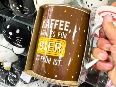 Villach, Avusturya - 17 Eylül 2025: Komik bira alıntısı olan kahverengi kupa canlı bir dükkanda kedi temalı oyuncaklarla çevrili