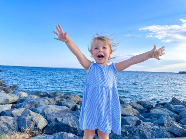Deniz kıyısında dikilen mavi elbiseli şirin küçük kız ellerini kaldırdı. Denizde bir tatil yaptığı için mutlu. Yüksek kalite fotoğraf