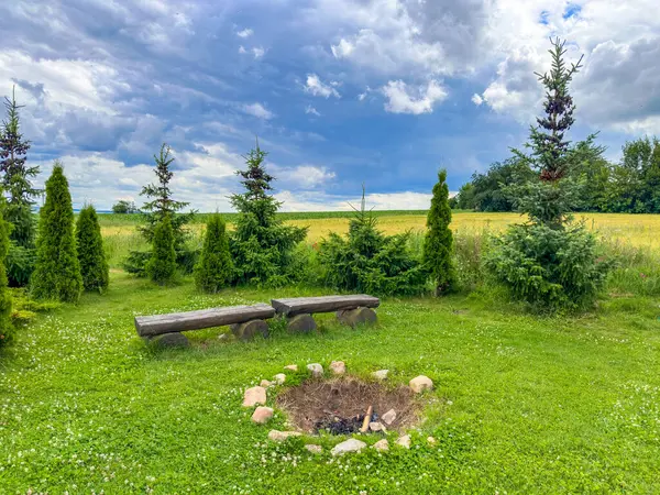Çimenli bir açık alanda kamp ateşi ve ahşap banklar. Ağaçlar ve kırsal bir çit açık hava ve kamp teması için mükemmel, rahat ve huzurlu bir ortam yaratır. Yüksek kalite fotoğraf
