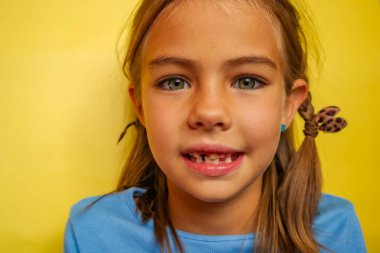 Dişleri dökülmüş, mavi tişörtlü, sarı arka planda gülümseyen genç bir kız. Kahverengi saçlı ve mavi gözlü. Bebek dişleri kalıcı dişlerle değiştiriliyor. Yüksek kalite fotoğraf