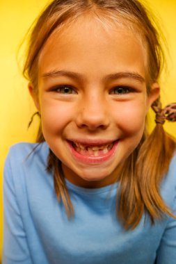 Dişleri dökülmüş, mavi tişörtlü, sarı arka planda gülümseyen genç bir kız. Kahverengi saçlı ve mavi gözlü. Bebek dişleri kalıcı dişlerle değiştiriliyor. Yüksek kalite fotoğraf