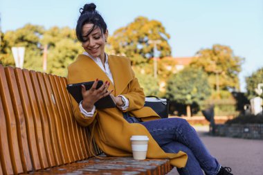 Parkta bankta oturan mutlu kadın tabletini okuyor ve kahve içiyor. Arka planda Tress, ceket ve kot pantolon giyiyor..