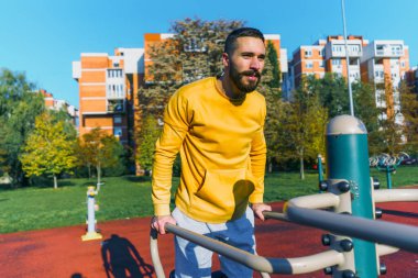 Genç sporcu güneşli bir günde açık hava spor salonunda barfiks çekiyor. Egzersizine odaklanıyor.