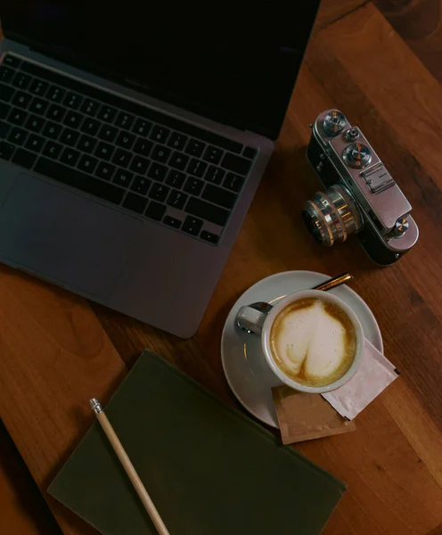 Photo of a cup of coffee with late art drawn on it, laptop notes and a vintage camera on the table next to the coffee cup