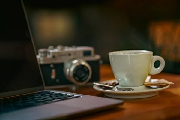 Photo of a cup of coffee with late art drawn on it, laptop notes and a vintage camera on the table next to the coffee cup