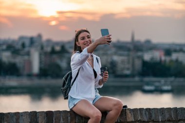 Eski kalenin tuğla çitlerinde oturan güzel bir turist kızı portresi. Arka planda şehir ve nehir ile fotoğraf çekmek için telefonunu kullanıyor..