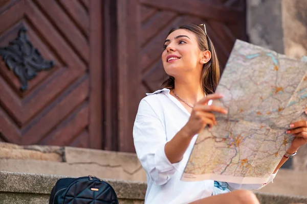 Kafkas kadın turist kağıt haritayla merdivenlerde oturuyor ve gündüz vakti güneşli şehir caddesine bakıyor.