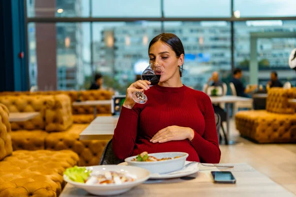Beautiful pregnant woman drinking wine. Young woman with glass of wine sitting at table in restaurant with a busy background